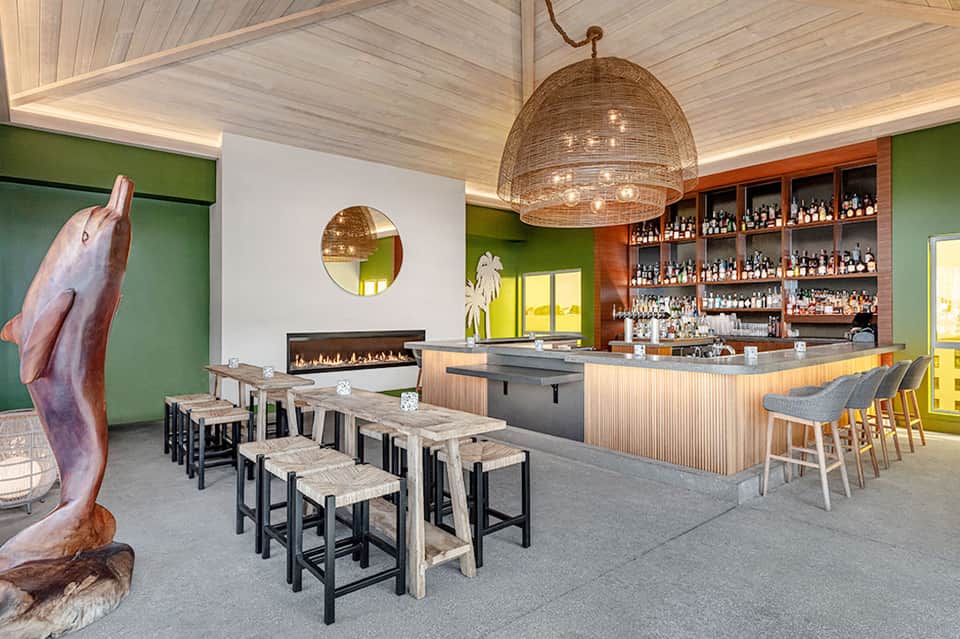 Modern hotel bar with green accent walls, wooden elements, fireplace, woven dome pendant, and wood seating