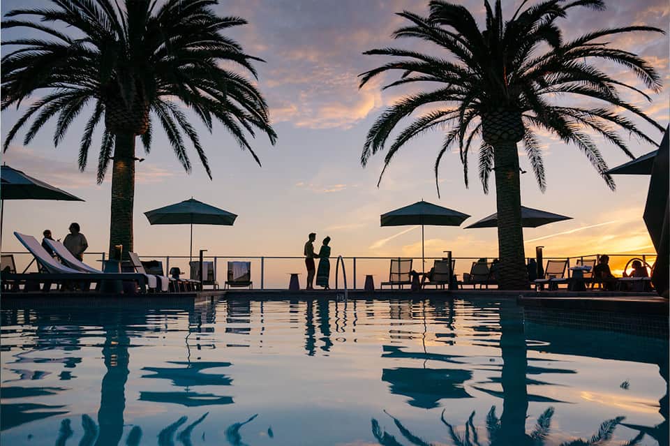 Rooftop pool at sunset with palm trees, lounge chairs, umbrellas, and silhouetted guests