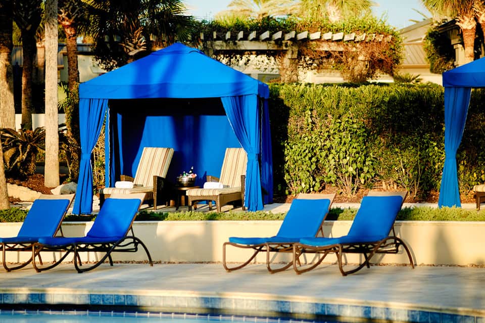 Outdoor poolside cabana with blue drapes and lounge chairs under palm trees at resort