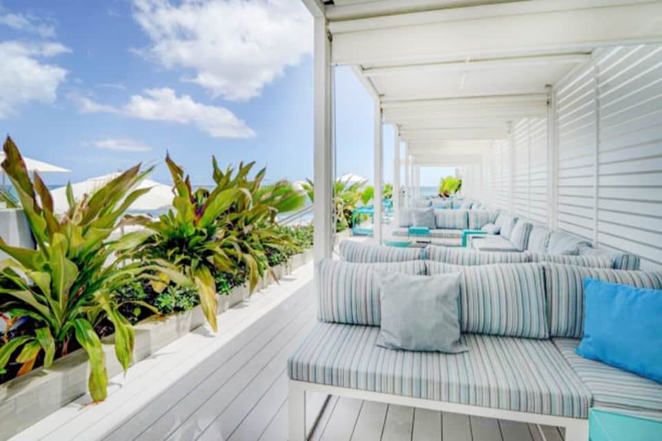 Covered beachside lounge area with striped cushioned seating, white pergola, and ocean horizon view