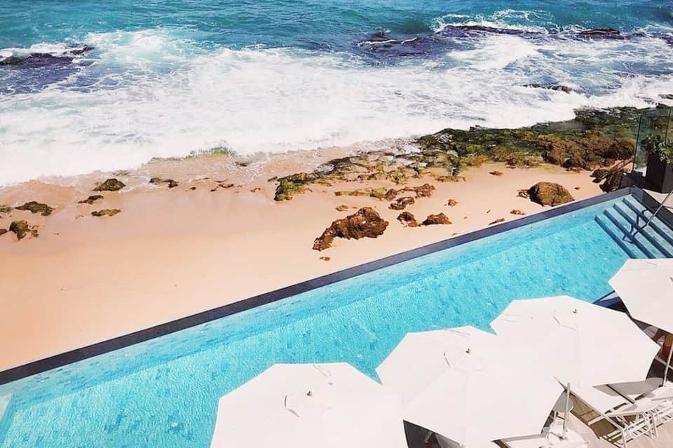 Oceanfront pool with white umbrellas overlooking beach, rocks, and turquoise sea
