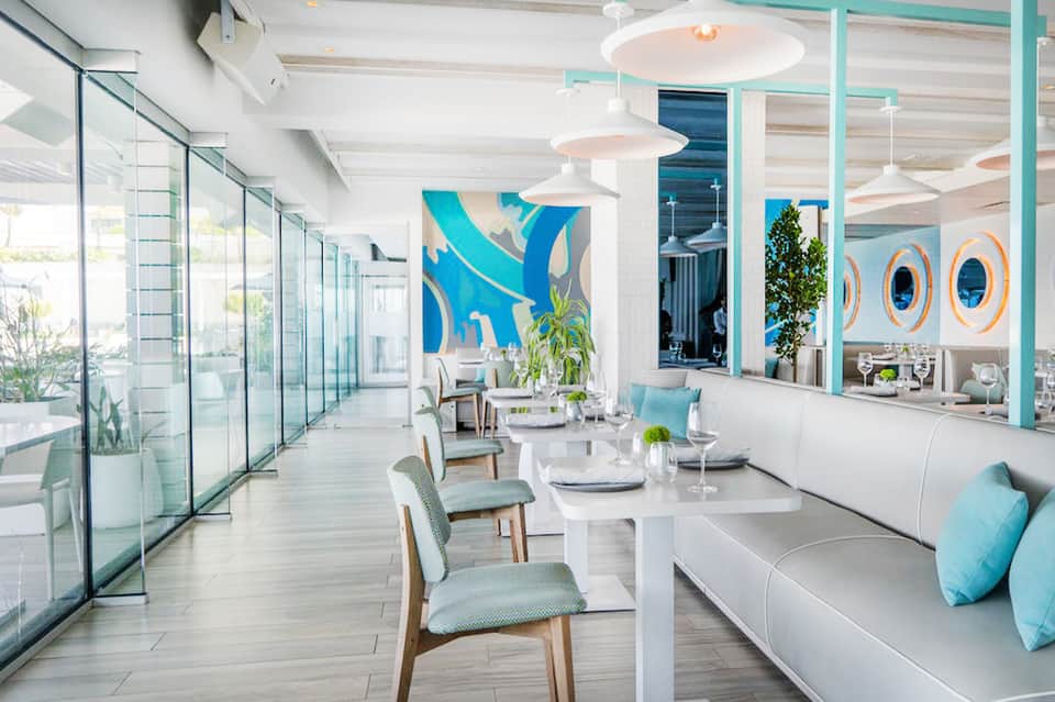 Modern restaurant interior with turquoise accents, white dining tables, glass walls, and coastal decor