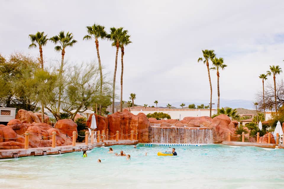 Resort pool with turquoise water, red rock features, waterfall, and palm trees under blue sky