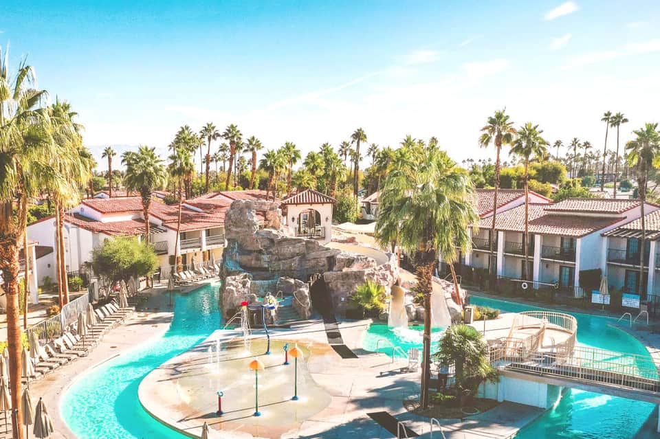 Tropical resort pool area with rock formations, palm trees, turquoise water, and Spanish architecture