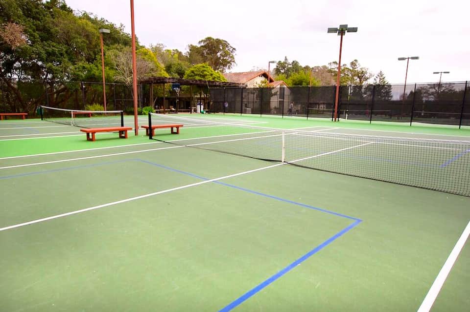 Outdoor tennis courts with green playing surfaces, benches, and tall light poles surrounded by trees