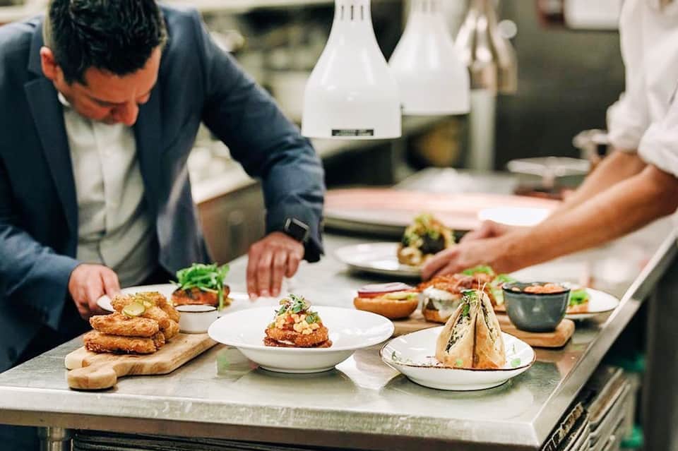 Chef plating gourmet dishes in professional kitchen with white pendant lights and stainless steel counters