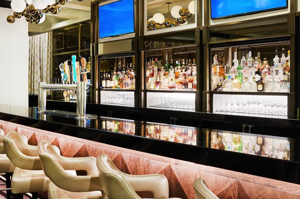 Upscale bar with extensive bottle display, brass fixtures, and cream-colored seating