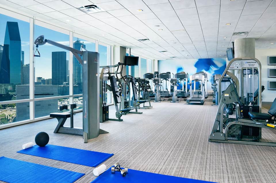 High-rise hotel fitness center with exercise equipment, blue yoga mats, and floor-to-ceiling windows overlooking city skyline