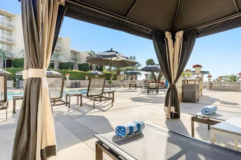 Covered outdoor seating pavilion with curtains overlooking resort pool and landscaping