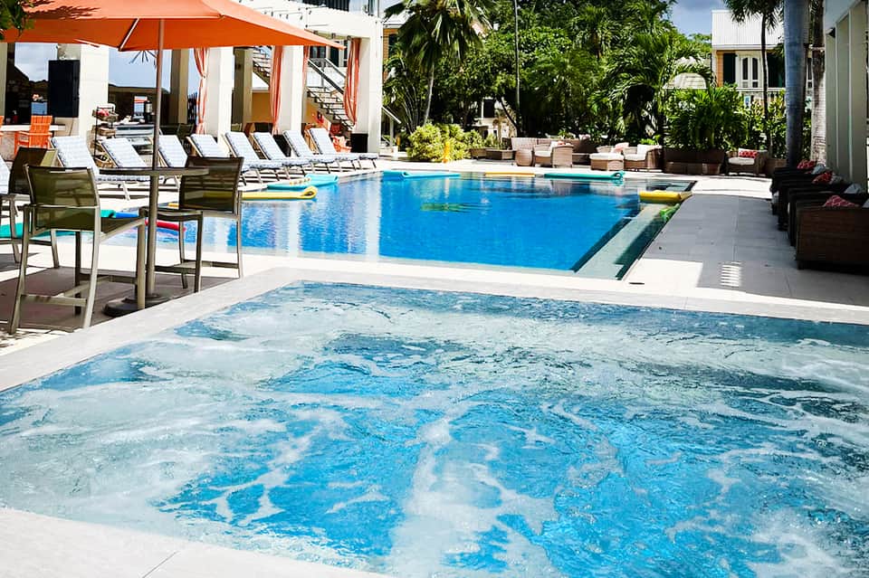 Resort pool area with hot tub, blue water, white loungers, orange umbrellas, and tropical trees