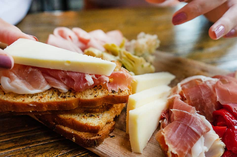 Hands assembling cured meats, cheese, and bread on a wooden serving board