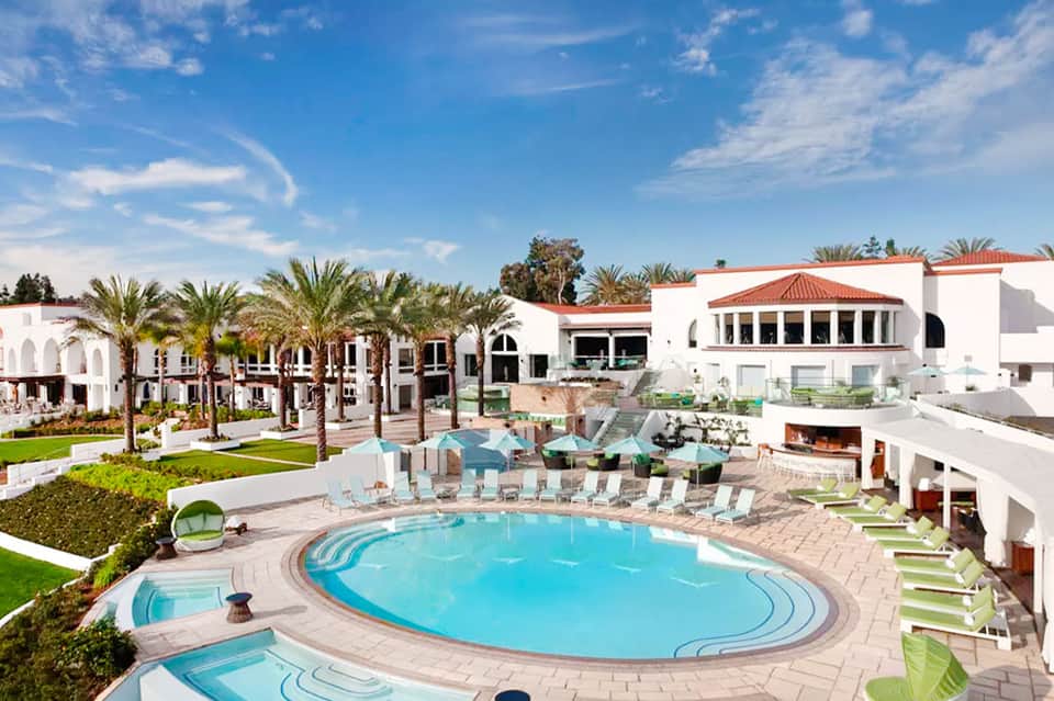 Resort courtyard with oval swimming pool, palm trees, white buildings, and lounge chairs