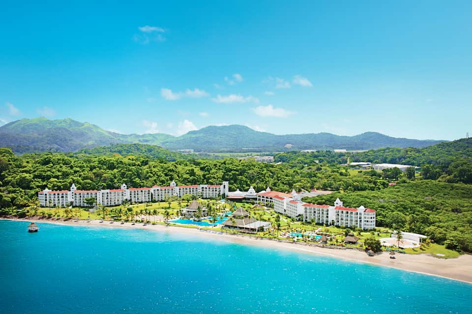 Aerial view of beachfront resort with white buildings, pool area, and mountains in background.
