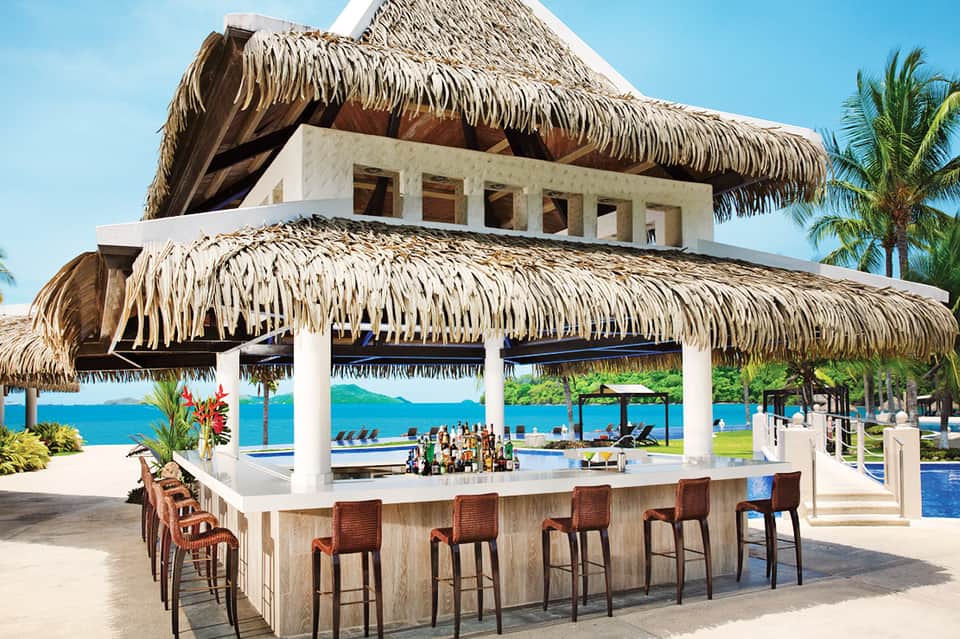 Beachfront bar with thatched roof, white columns, pool view, and blue ocean horizon