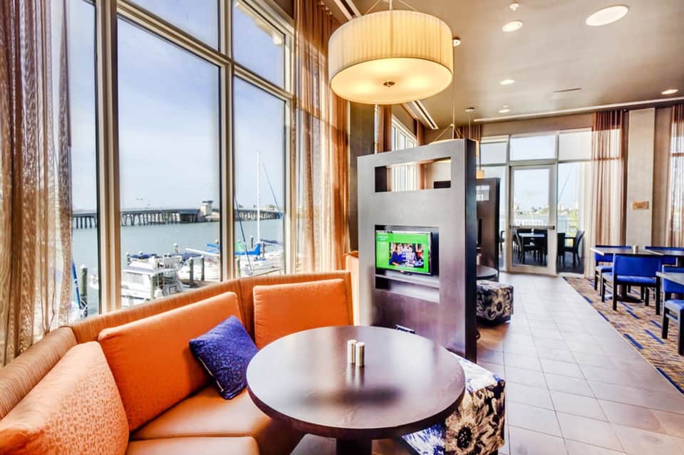 Modern hotel suite lounge with orange seating, water views through floor-to-ceiling windows, and fireplace