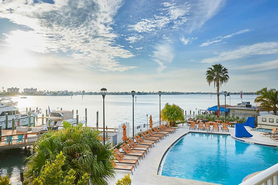 Resort pool overlooking bay with lounge chairs, palm trees, and boats