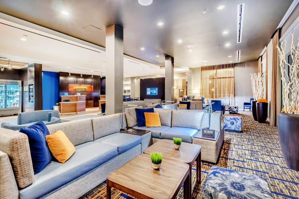 Modern hotel lobby lounge with gray couches, blue and orange pillows, wood tables and bar