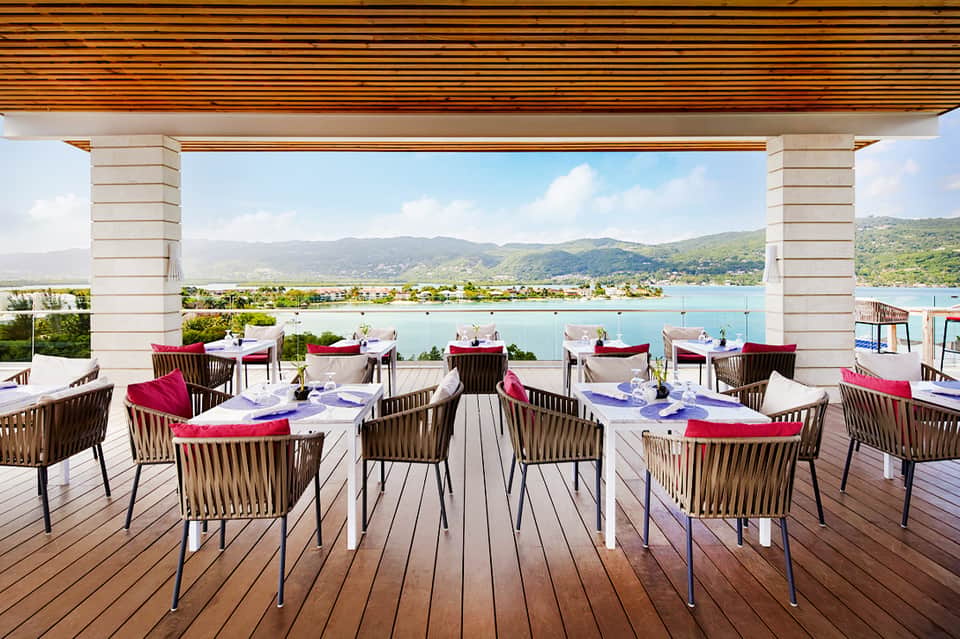 Waterfront dining deck with white tables, wooden chairs, red cushions, and mountain lake views