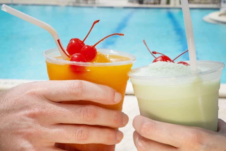 Hands holding colorful cocktails with cherries by pool, bright sunny day with turquoise water visible