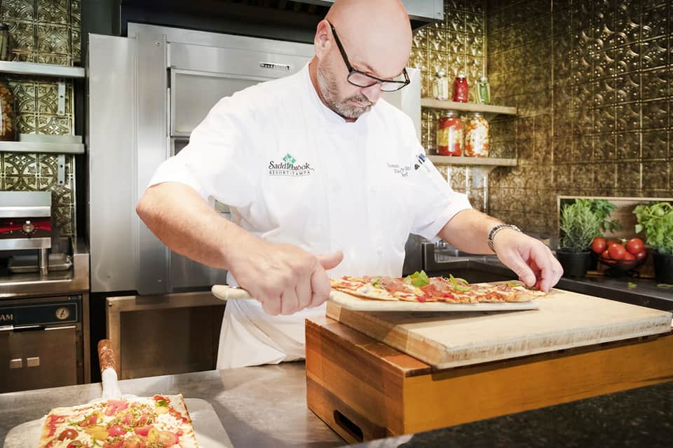 Chef in white uniform preparing pizza in modern kitchen with gold patterned backsplash and shelving