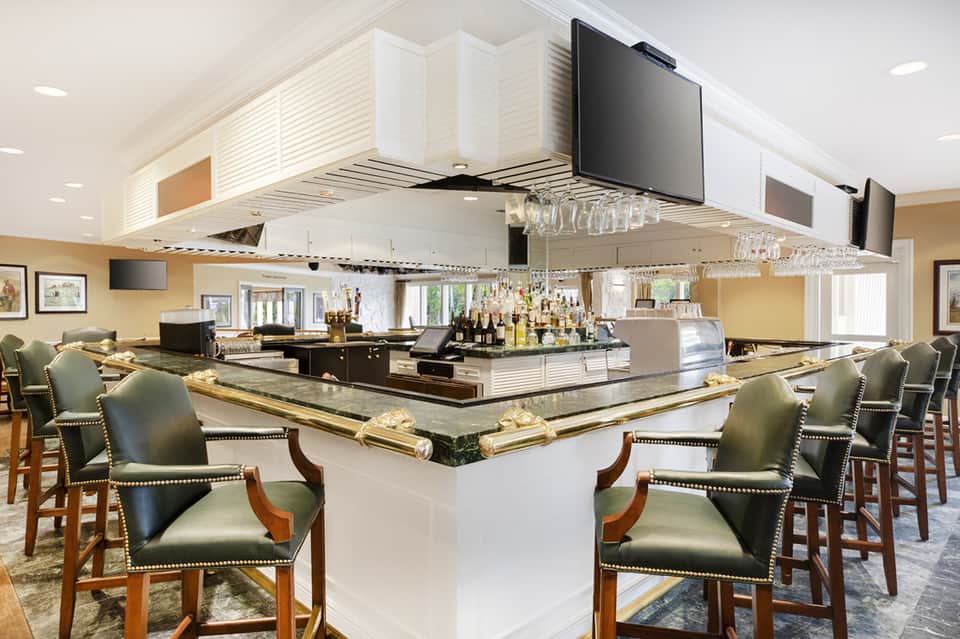 Upscale indoor bar with central island counter, green leather seating, and multiple screens