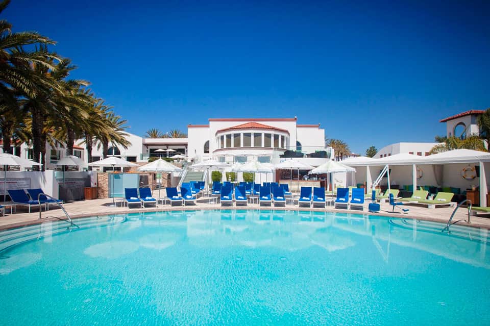 Resort pool with turquoise water, blue lounge chairs, white umbrellas, and white building with red tile roof