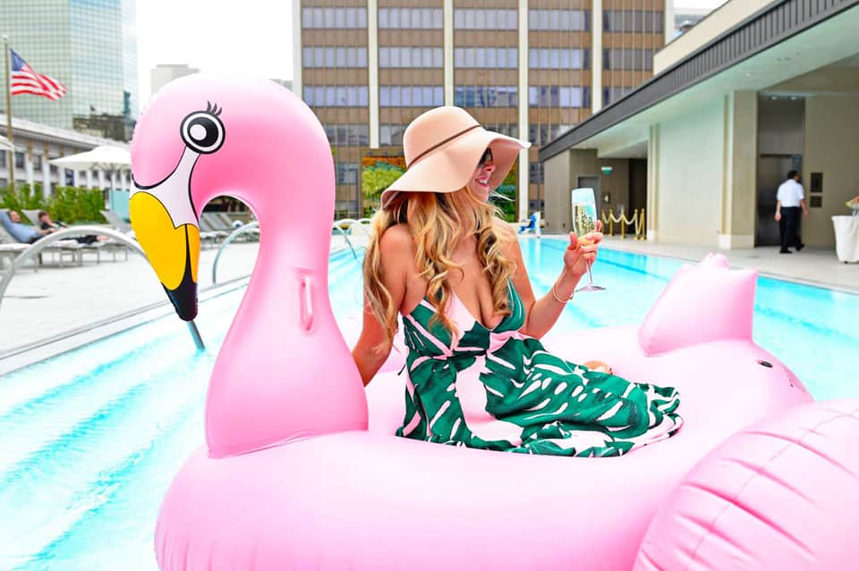 Woman on pink flamingo pool float at urban hotel pool wearing green tropical print swimsuit and peach hat