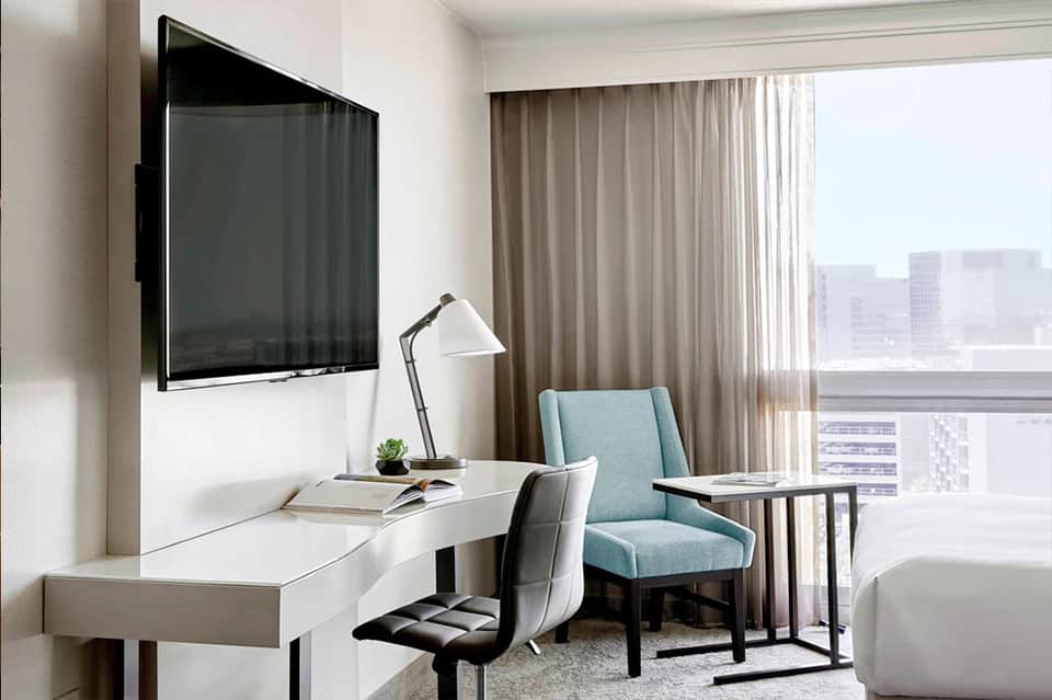 Modern hotel room with desk, blue accent chair, wall-mounted TV, and city skyline view