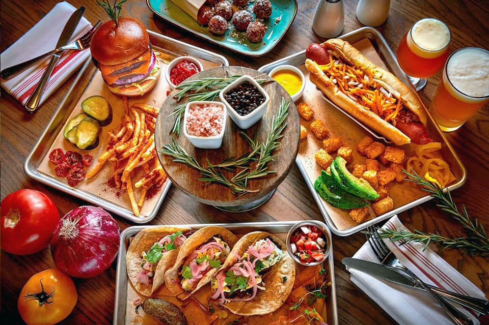 Overhead view of gourmet food spread with burgers, hot dogs, tacos, and beverages