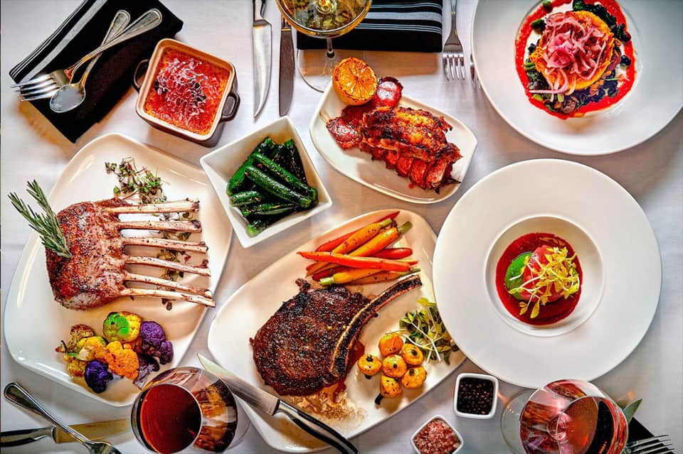 Fine dining spread with grilled steak, seafood, soup, and colorful vegetables on white plates