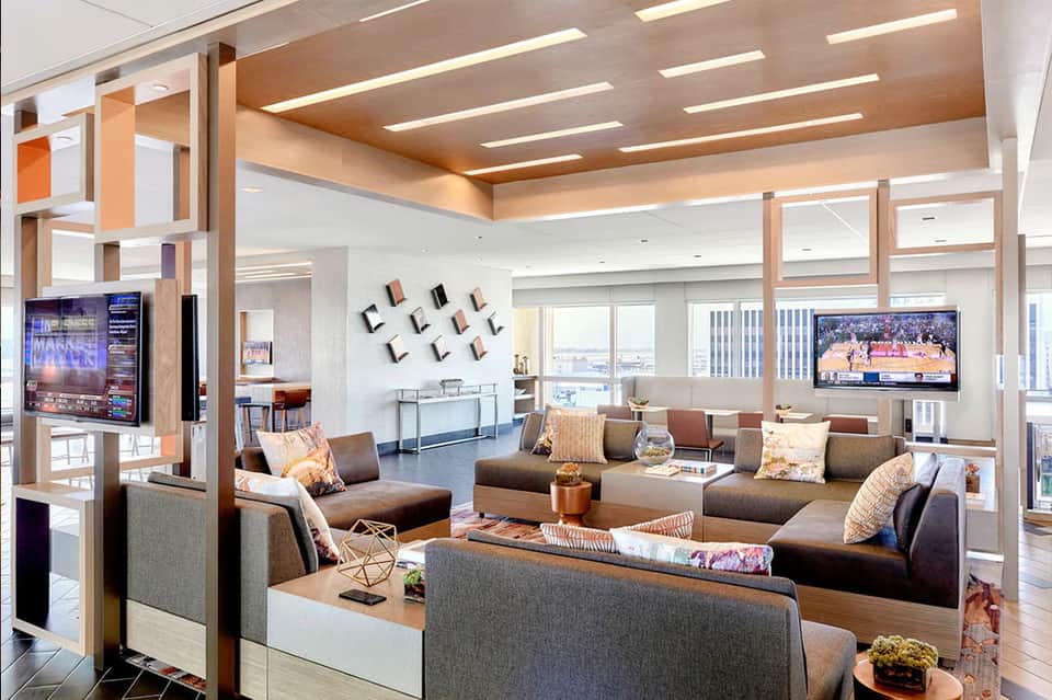 Modern hotel lounge with wood ceiling beams, brown sectional seating, floor-to-ceiling windows with city views, and wall-mounted TV