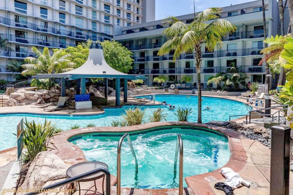Resort courtyard with sparkling blue pool, hot tub, tropical landscaping, and multi-story hotel buildings