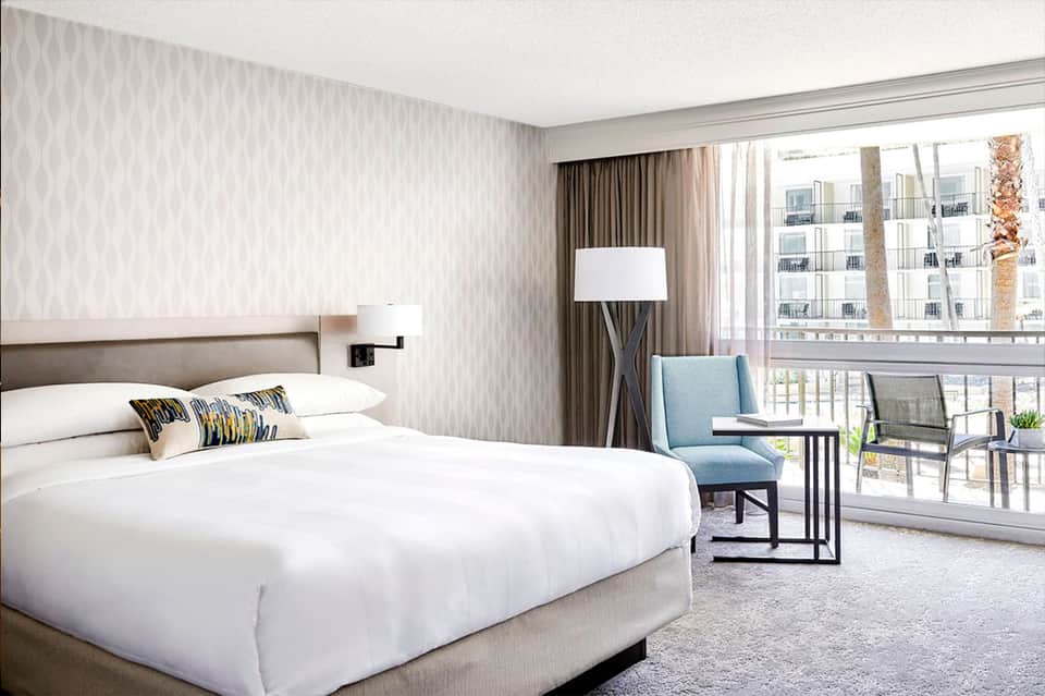 Spacious hotel bedroom with king bed, seating area, balcony overlooking palm trees and cityscape