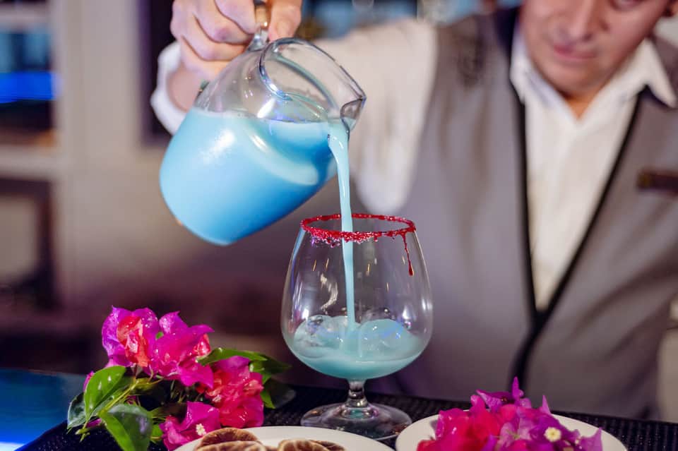 Bartender pouring vibrant blue cocktail into red-rimmed glass with pink flowers on table