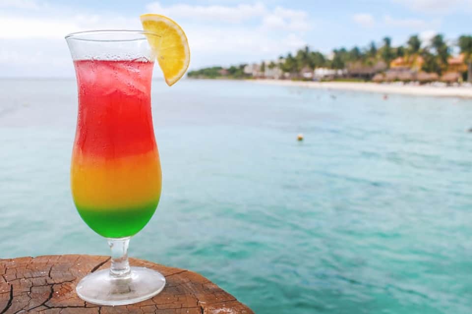 Rainbow layered cocktail with lemon slice on a wooden dock overlooking tropical beach
