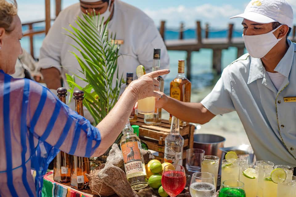 Beachfront bar service with bartender preparing tropical cocktails overlooking ocean with palm trees and bottles