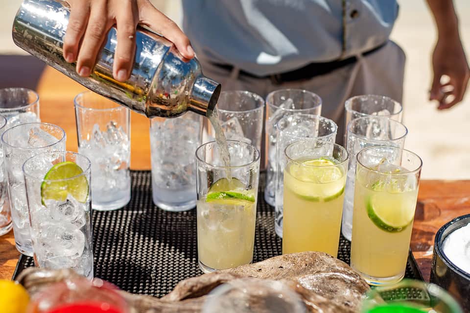 Server pouring tequila into multiple glasses with lime wedges on black surface