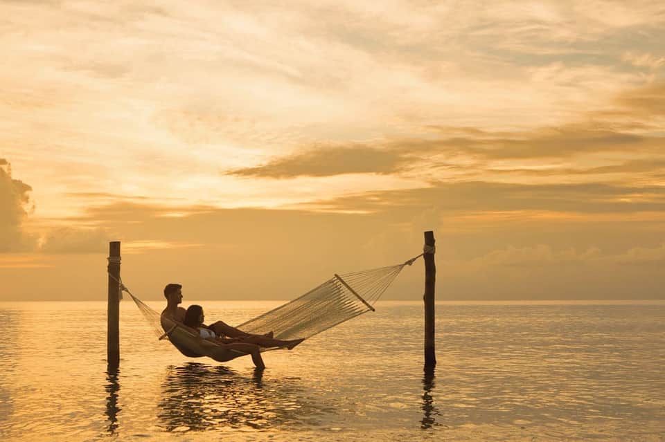 Couple relaxing in hammock at sunset over calm ocean water during golden hour