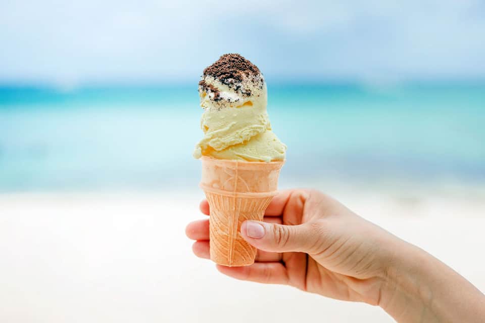 Hand holding vanilla ice cream cone with sprinkles against white sand beach backdrop