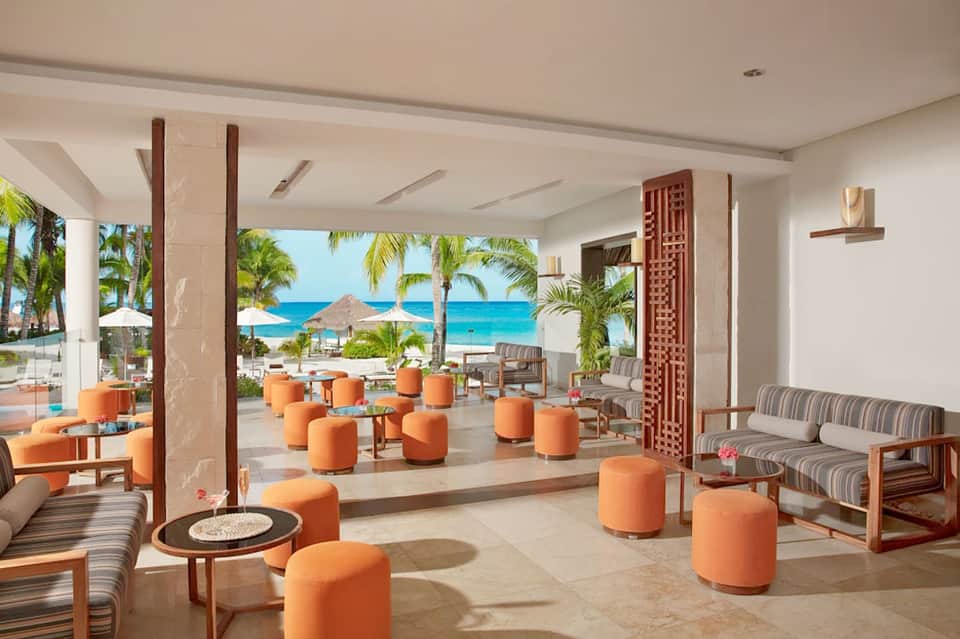 Bright resort lounge with orange accent seating, beach view, palm trees, thatched umbrella, and tropical plants
