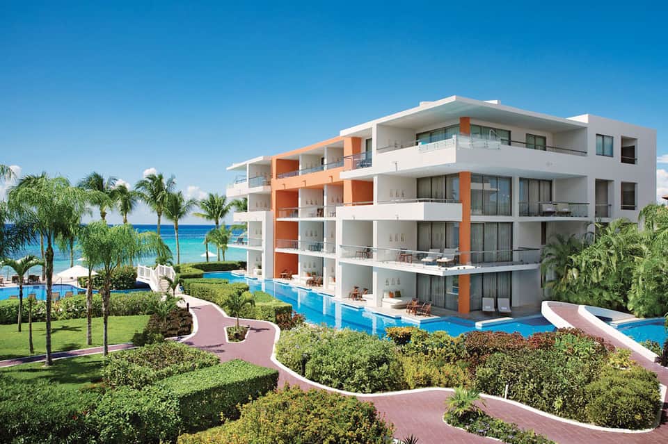 Modern beachfront resort building with white and orange architecture, multiple balconies, pools, and tropical landscaping