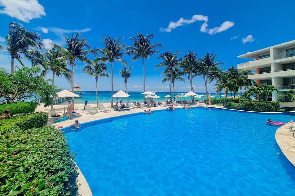 Beachfront resort pool with ocean view, palm trees, white umbrellas, and sandy beach beyond