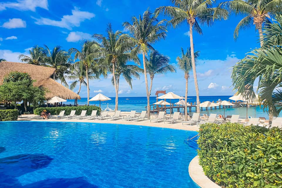 Luxury beachfront resort pool with palm trees, white umbrellas, ocean view, and pier