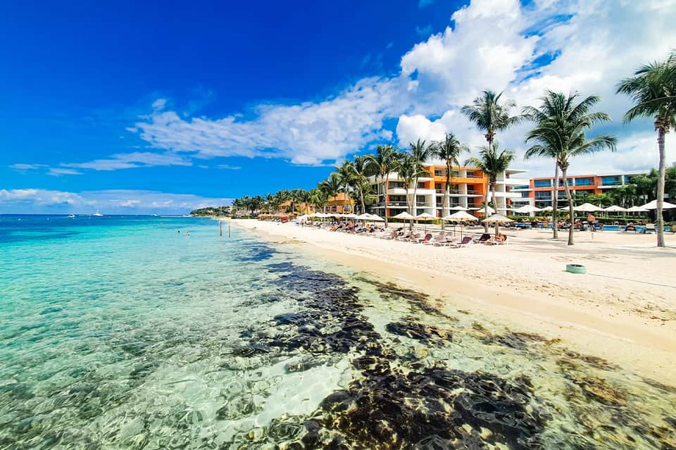 Tropical beach resort with turquoise water, sandy beach, colorful buildings, palm trees, and lounge chairs