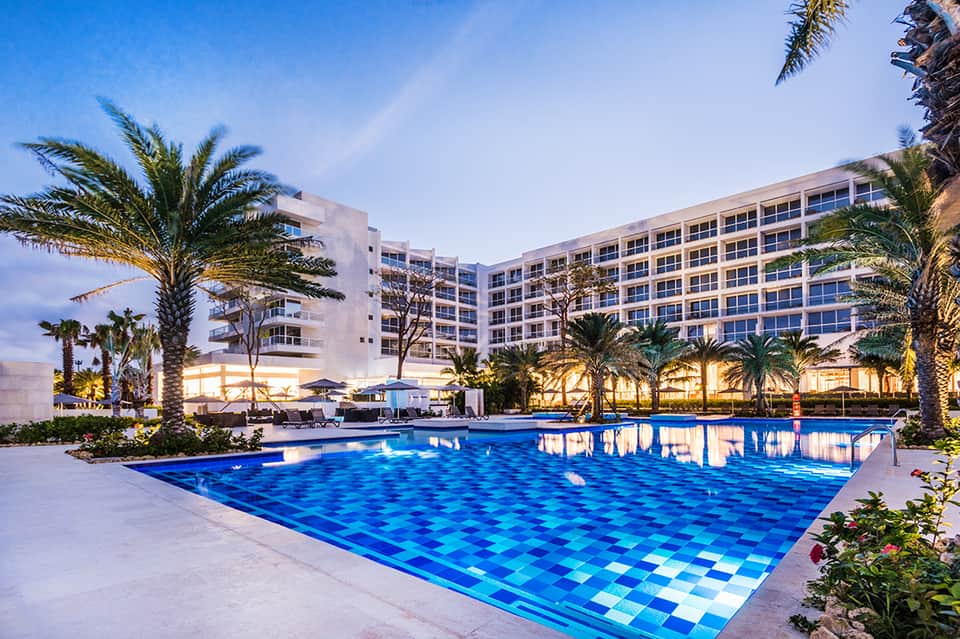 Modern resort at dusk with illuminated pool, palm trees, and multi-story building
