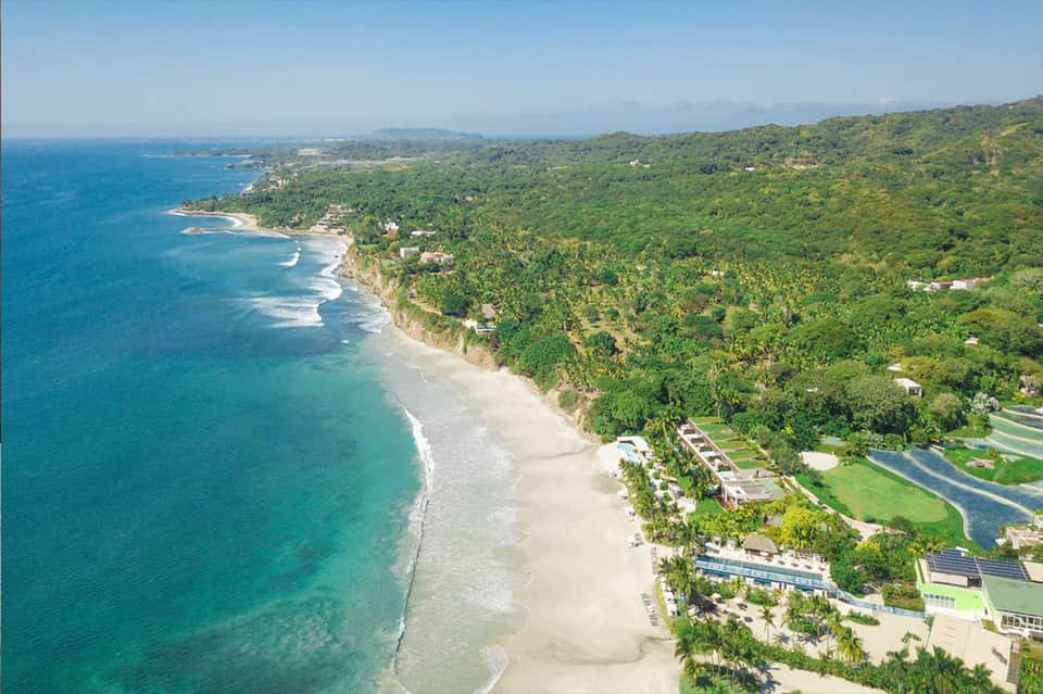 Aerial view of pristine beach with turquoise waters, lush green hillside, and luxury coastal development