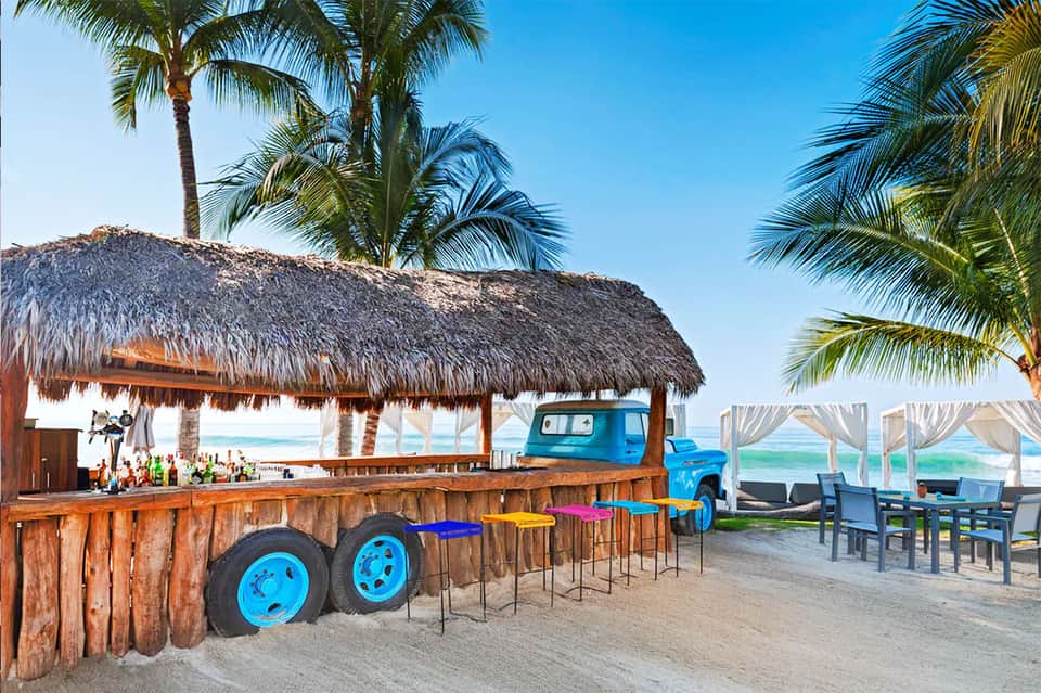 Beachfront tiki bar with thatch roof, blue truck decor, colorful stools, and ocean view