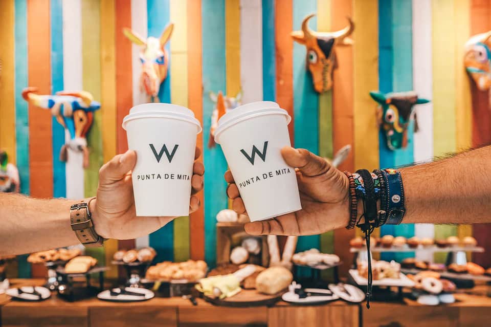 Two white coffee cups with W Punta de Mita logo held in front of colorful striped wall