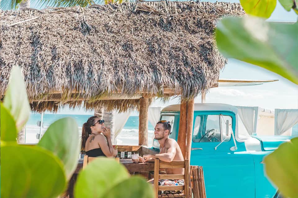 Couple dining under thatched-roof beach cabana with vintage truck
