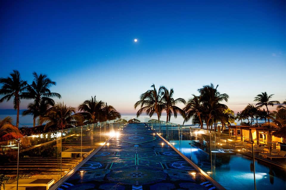 Illuminated boardwalk over turquoise water at dusk with palm trees and resort buildings